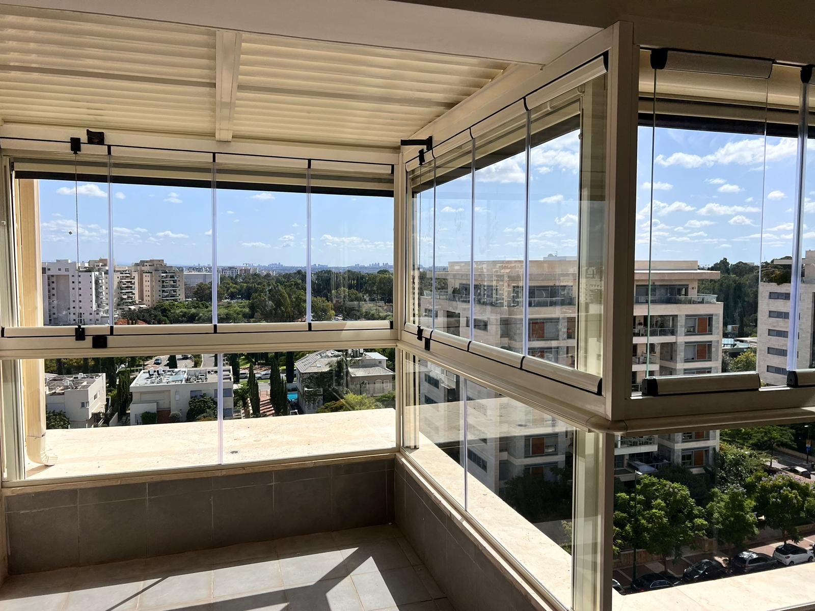 סגירת זכוכית למרפסת פינתית בפרופילים בגוון שמנת, נוף עירוני ירוק