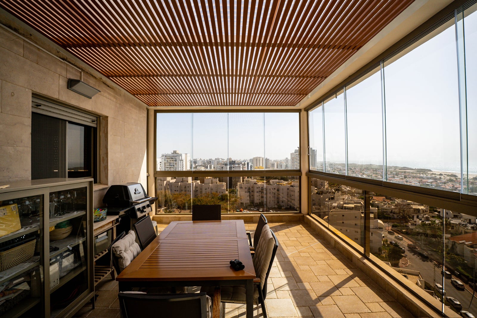 מרפסת סגורה בזכוכית עם תקרת פרגולה בגוון עץ, שולחן אוכל, גריל ונוף לעיר ולים