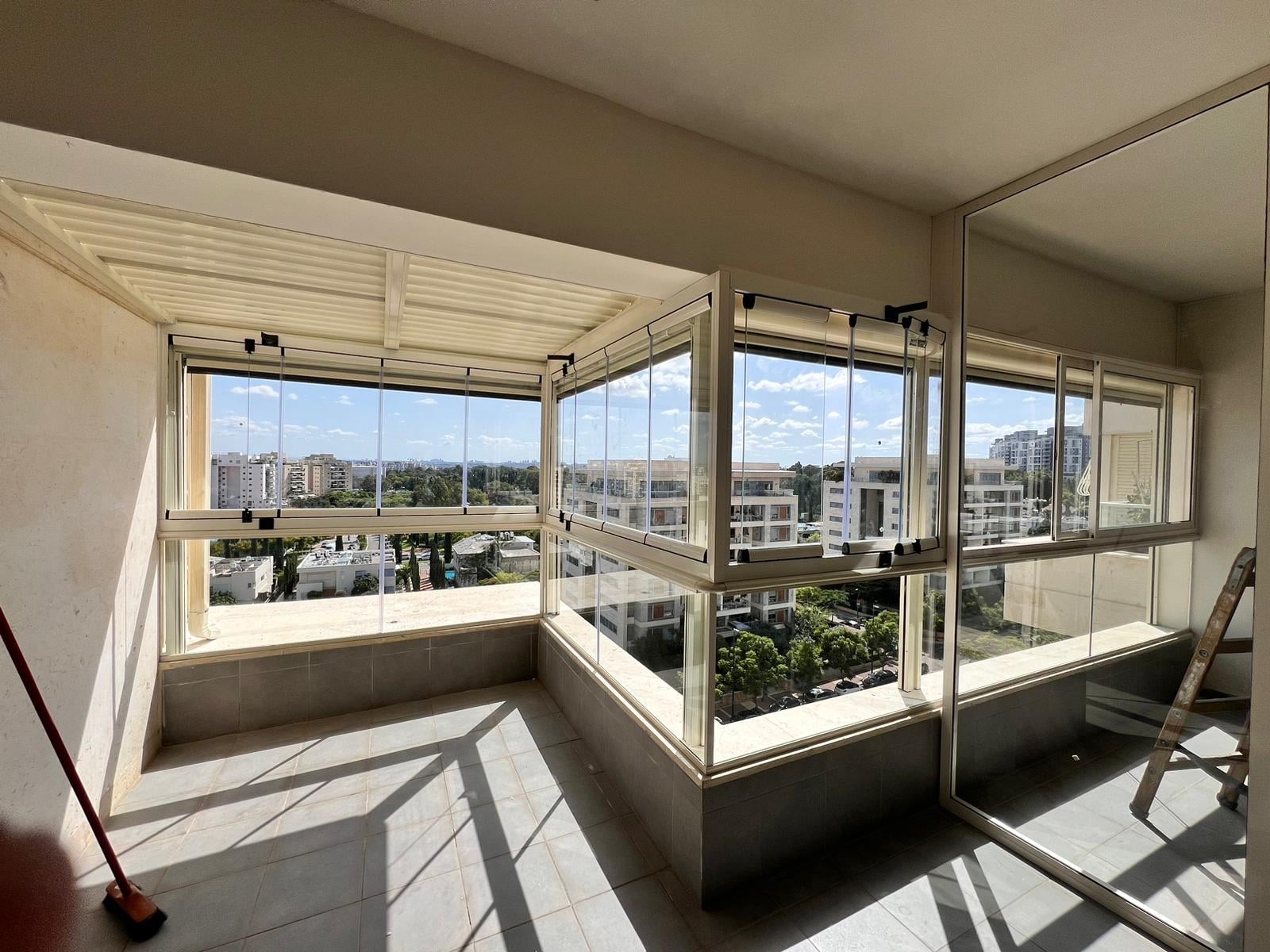 מרפסת פינתית סגורה בזכוכית במסגרות שמנת, תקרת לוברים לבנה ונוף עירוני