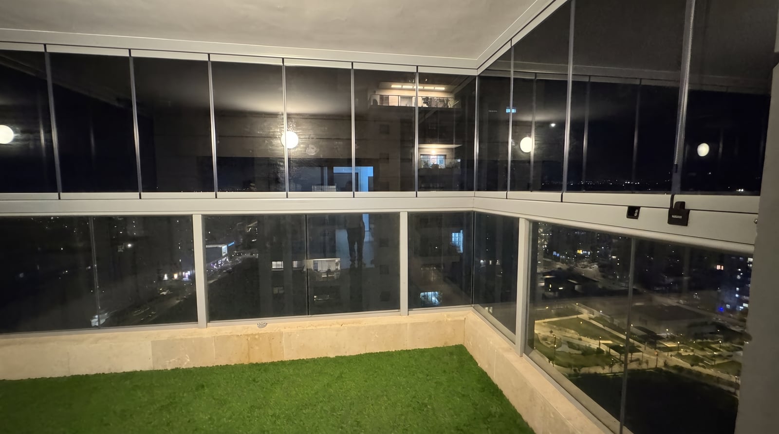 מרפסת סגורה בזכוכית בלילה, פרופילים אפורים בהירים ודשא סינתטי על הרצפה