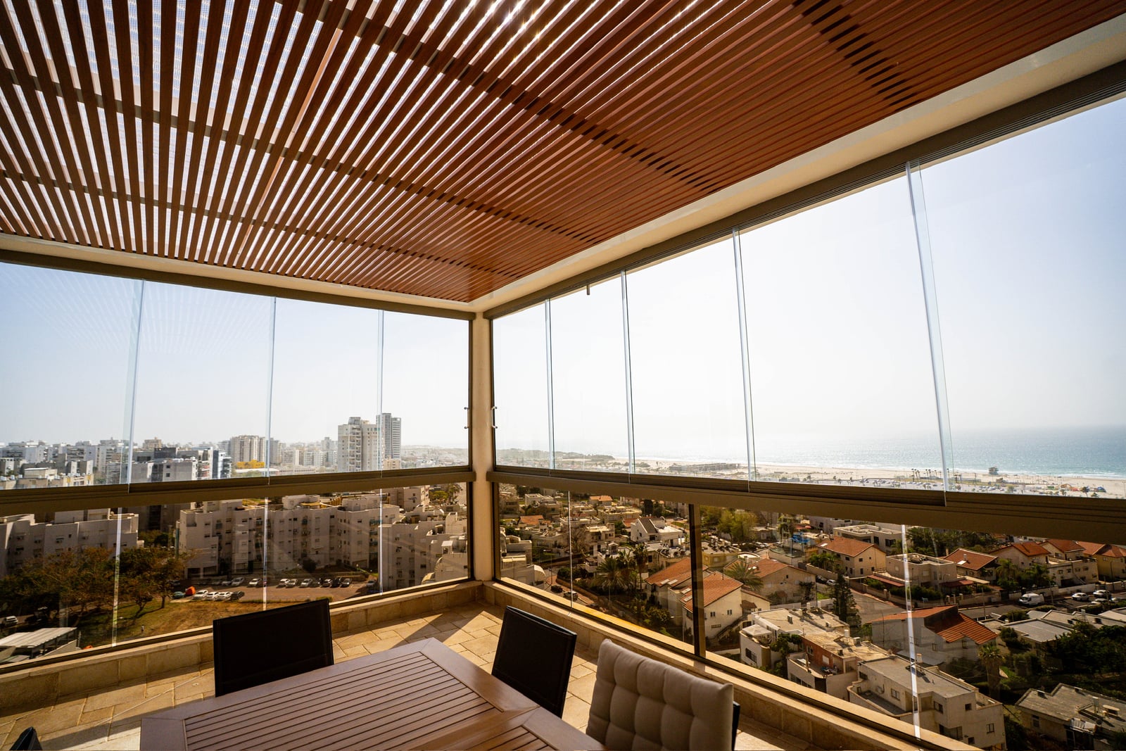 מרפסת פינתית סגורה בזכוכית מתחת לפרגולה דמוי עץ, מול חוף הים