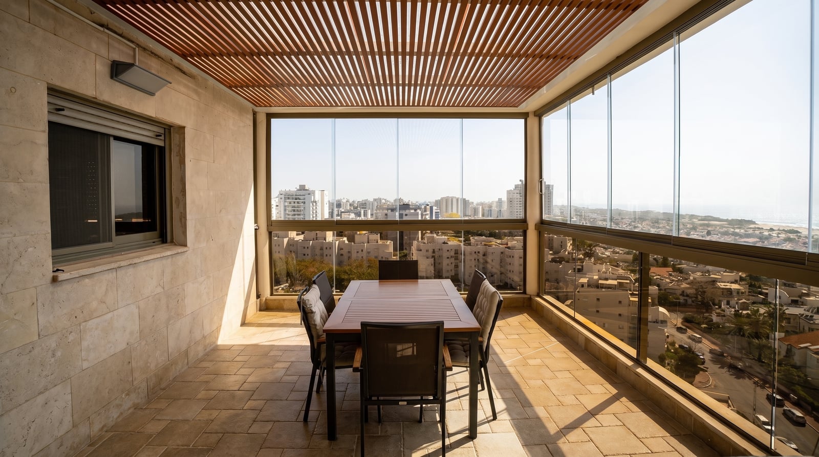מרפסת עם סגירת זכוכית ותקרת פרגולה בגוון עץ, פינת אוכל ונוף עירוני