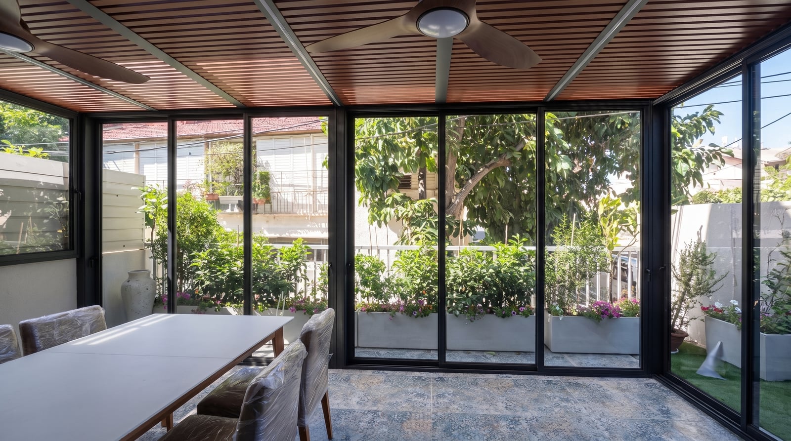 חדר זכוכית במסגרות שחורות עם תקרת פרגולה בגוון עץ, מאווררים ופינת אוכל