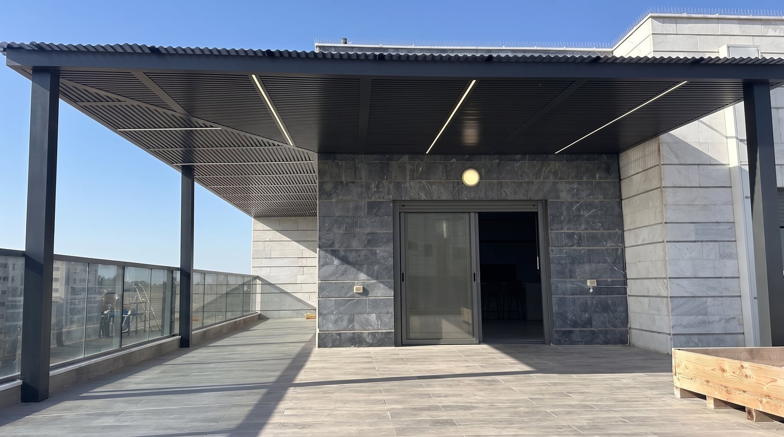 פרגולה אפורה כהה משלבים עם פסי לד במרפסת גג, מול קיר אבן