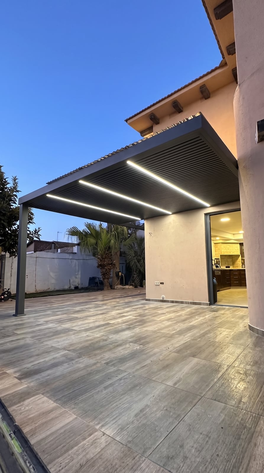 פרגולת אלומיניום אנתרציט עם פסי לד מעל פטיו מרוצף ליד הבית, בשעת דמדומים