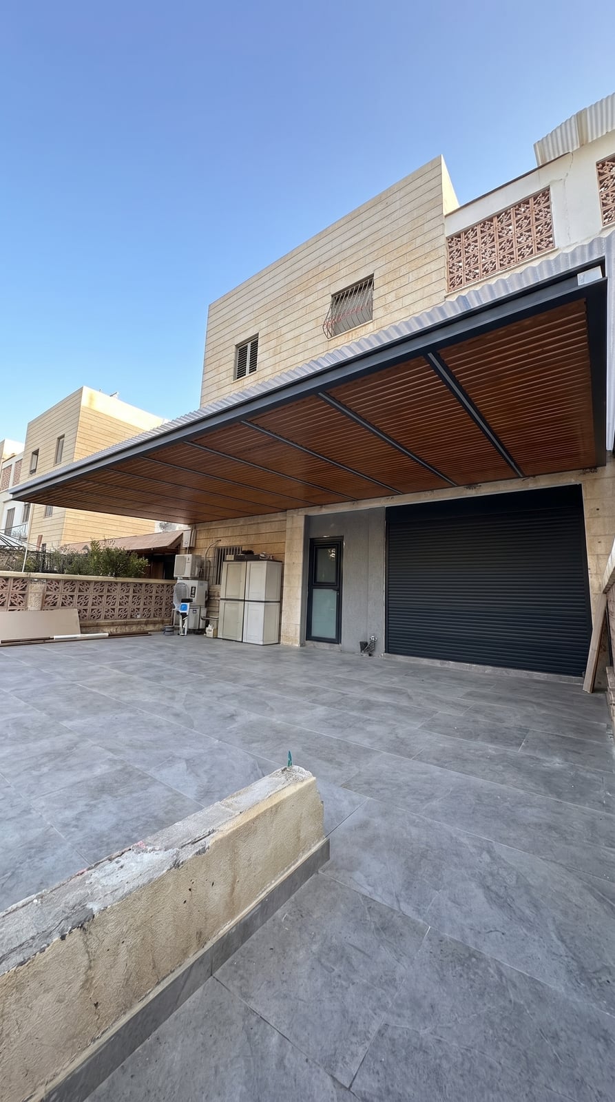פרגולה גדולה בגוון עץ עם מסגרת כהה מעל חצר כניסה של בית אבן