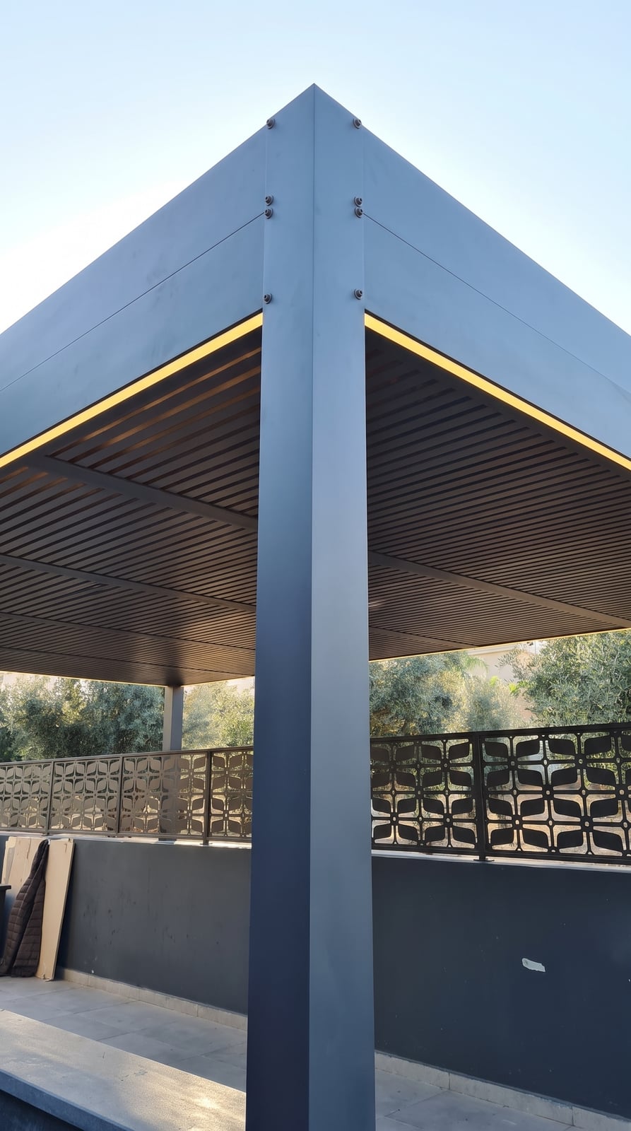 פינת פרגולה באפור כהה עם תאורת לד היקפית ותקרת שלבים, ליד מעקה דקורטיבי