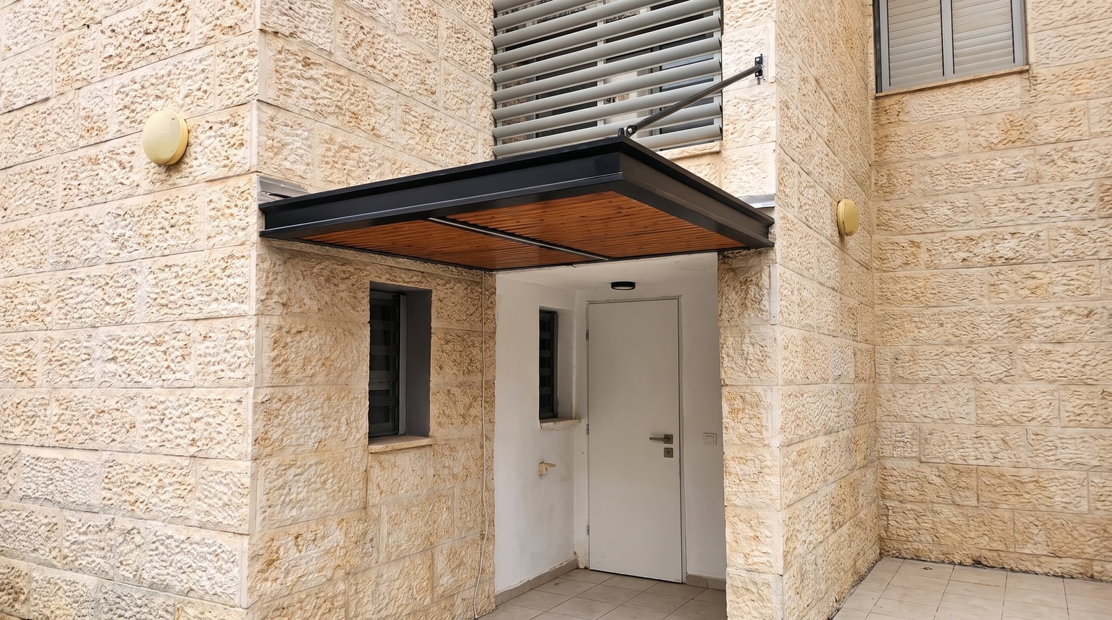 גגון בגוון עץ עם מסגרת שחורה מעל דלת כניסה בבניין מחופה אבן
