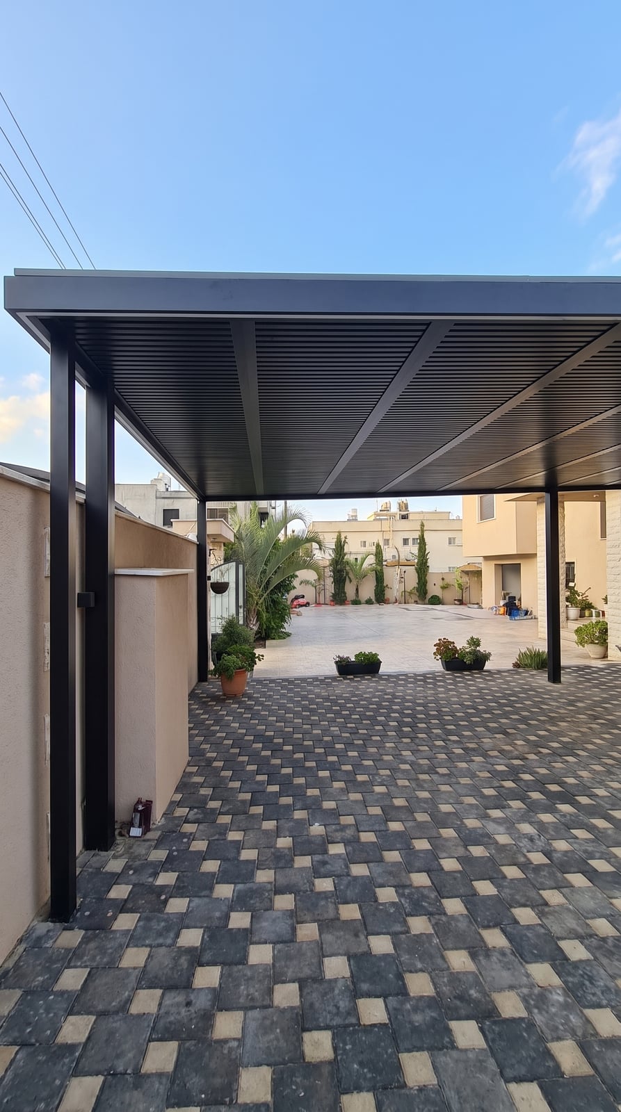 פרגולה שחורה עם תקרת שלבים מעל רחבה מרוצפת באבנים משתלבות