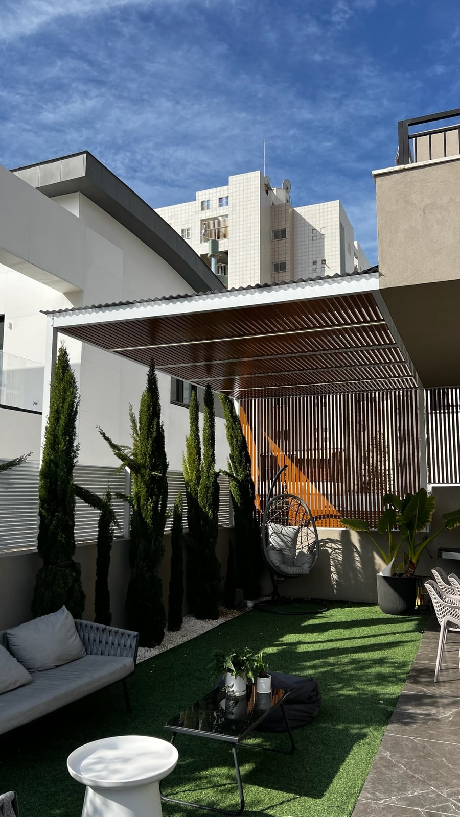 פרגולה עם מסגרת לבנה ושלבים בגוון עץ מעל חצר עם דשא סינתטי וכורסה תלויה