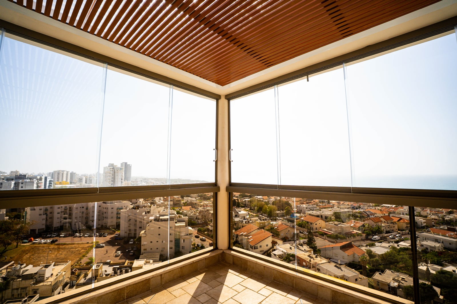 מרפסת עם סגירת זכוכית ותקרת פרגולה בגוון עץ, נוף עירוני וים